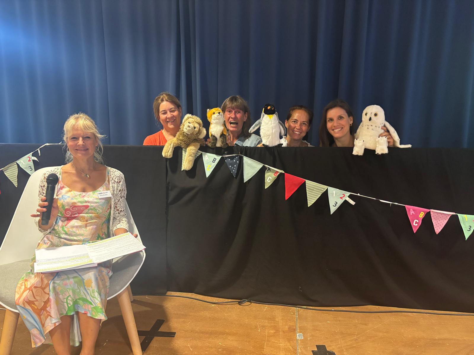 Eine Frau sitzt mit Mikrofon und Unterlagen auf einem Stuhl vor einem Vorhang, hinter der mehrere Personen mit Stofftieren.